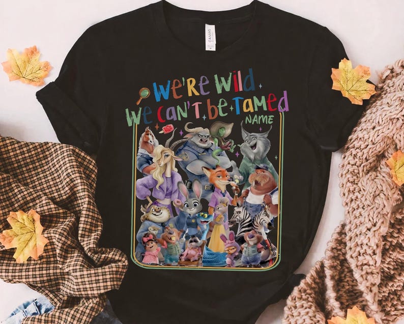 May include: Black t-shirt featuring a colorful graphic of various cartoon characters. The text "We're Wild, We Can't Be Tamed" is displayed above the characters, with a space for a name below. The design is outlined in a gold border.
