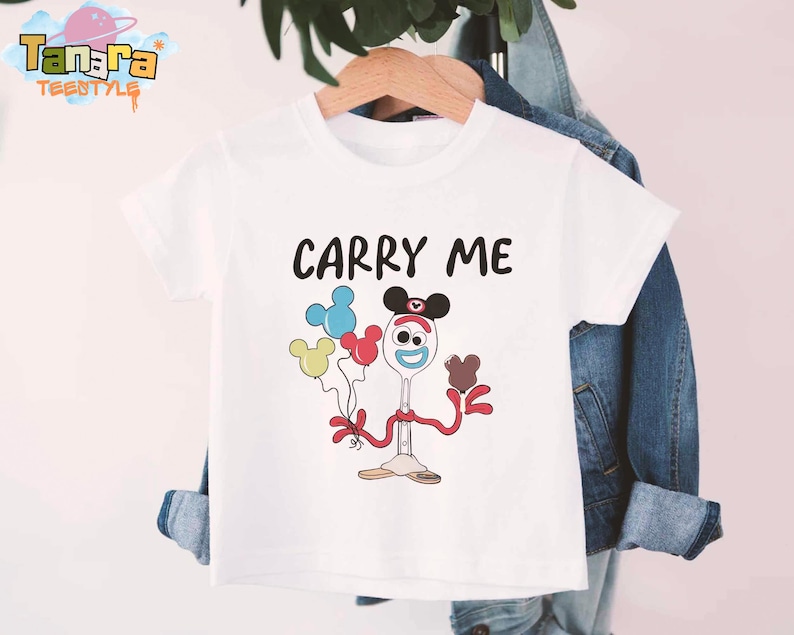 K&ouml;nnte beinhalten: Wei&szlig;es T-Shirt mit dem Text "CARRY ME" in Schwarz. Das Shirt zeigt eine Zeichentrickfigur, die Luftballons h&auml;lt. Die Figur ist ein L&ouml;ffel mit Mickey Mouse-Ohren und roten Armen und Beinen. Die Ballons sind blau, gelb und rot.