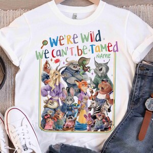 May include: White t-shirt featuring a colorful graphic of various cartoon animal characters. The text "WE'RE WILD WE CAN'T BE TAMED" is printed above the characters. The shirt is paired with blue jeans, white sneakers, and a watch.