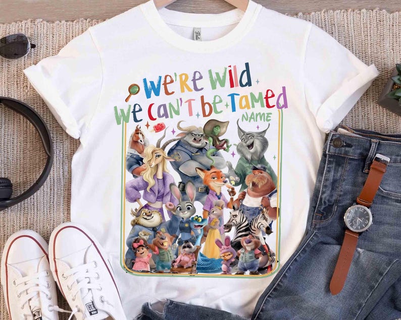 May include: White t-shirt featuring a colorful graphic of various cartoon animal characters. The text "WE'RE WILD WE CAN'T BE TAMED" is printed above the characters. The shirt is paired with blue jeans, white sneakers, and a watch.