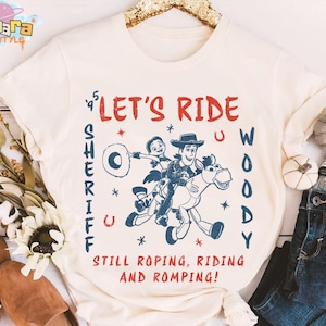 May include: Cream-colored t-shirt featuring a graphic of Woody and a friend riding a horse, with the text "LET'S RIDE WOODY SHERIFF" in red and blue. The shirt also includes the phrase "STILL ROPING, RIDING AND ROMPING!"