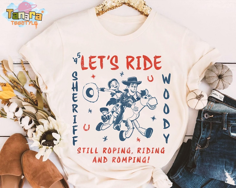 May include: Cream-colored t-shirt featuring a graphic of Woody and a friend riding a horse, with the text "LET'S RIDE WOODY SHERIFF" in red and blue. The shirt also includes the phrase "STILL ROPING, RIDING AND ROMPING!"