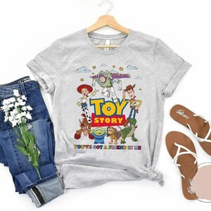 May include: A light gray t-shirt featuring the Toy Story characters with the words "TOY STORY" in large, colorful letters. The shirt also has the phrase "YOU'VE GOT A FRIEND IN ME". Blue jeans and brown sandals are also in the image.