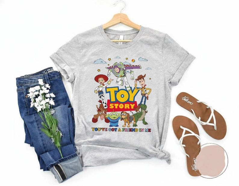 May include: A light gray t-shirt featuring the Toy Story characters with the words "TOY STORY" in large, colorful letters. The shirt also has the phrase "YOU'VE GOT A FRIEND IN ME". Blue jeans and brown sandals are also in the image.