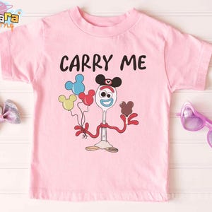 K&ouml;nnte beinhalten: Ein hellrosa T-Shirt mit dem schwarzen Schriftzug "CARRY ME". Darunter befindet sich eine Cartoonfigur, die Luftballons und ein Schokoladenherz h&auml;lt. Die Figur ist ein L&ouml;ffel mit Gesicht und Mickey Mouse Ohren.