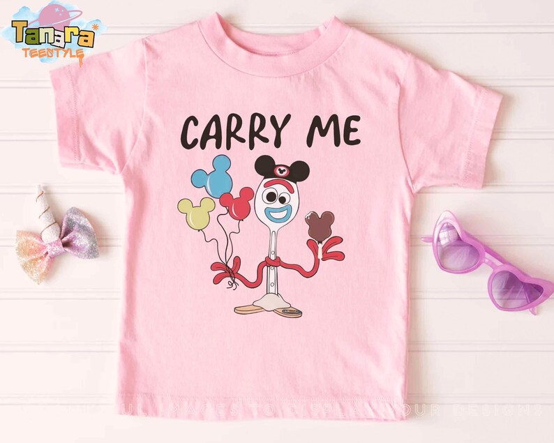 K&ouml;nnte beinhalten: Ein hellrosa T-Shirt mit dem schwarzen Schriftzug "CARRY ME". Darunter befindet sich eine Cartoonfigur, die Luftballons und ein Schokoladenherz h&auml;lt. Die Figur ist ein L&ouml;ffel mit Gesicht und Mickey Mouse Ohren.