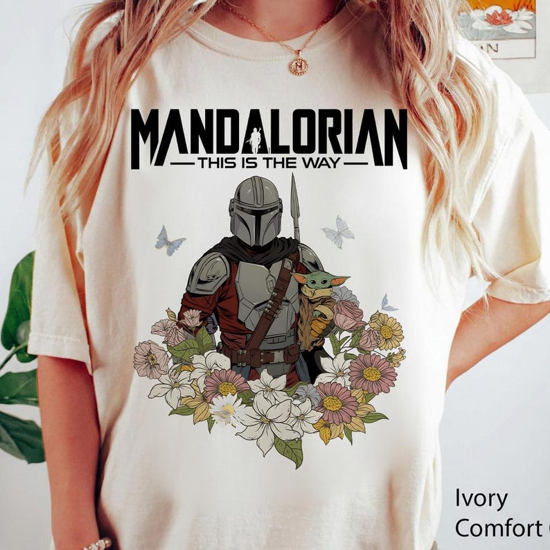 Puede incluir: Camiseta color marfil con un dise&ntilde;o del Mandaloriano sosteniendo a Grogu, rodeado de flores y mariposas. El texto "MANDALORIAN - THIS IS THE WAY" est&aacute; impreso encima del dise&ntilde;o. Corte c&oacute;modo.