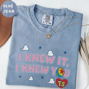 May include: A blue jean colored t-shirt featuring the text I KNEW IT, I KNEW YOU in pink letters. The design includes white clouds, blue stars, a colorful ball graphic, and a red heart with the yellow letters TS inside. A silver necklace is visible.