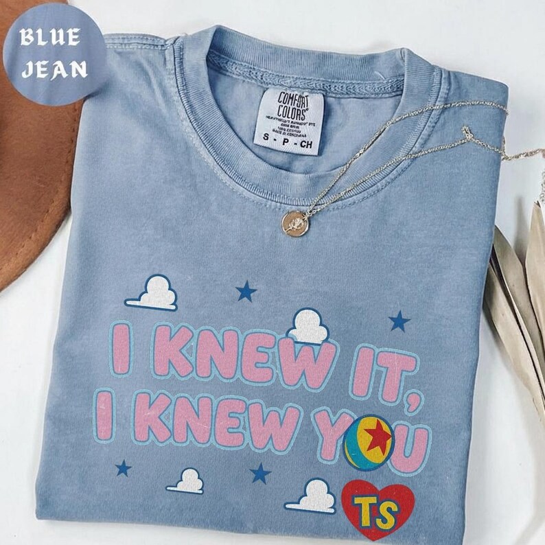 May include: A blue jean colored t-shirt featuring the text I KNEW IT, I KNEW YOU in pink letters. The design includes white clouds, blue stars, a colorful ball graphic, and a red heart with the yellow letters TS inside. A silver necklace is visible.