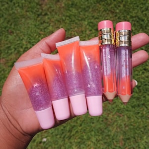 Peut inclure: Une collection de tubes de gloss à lèvres de différents modèles. Certains tubes ont la forme de crayons avec des gommes roses et des détails dorés. Les autres tubes sont transparents avec des paillettes orange et violettes. Les tubes de gloss ont des bouchons roses.