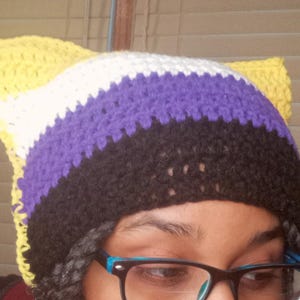 May include: A handmade crochet hat with cat ears, featuring the non-binary pride flag colors: yellow, white, purple, and black. The hat is visible from the forehead up, with the ears standing up. The hat is a unique accessory.