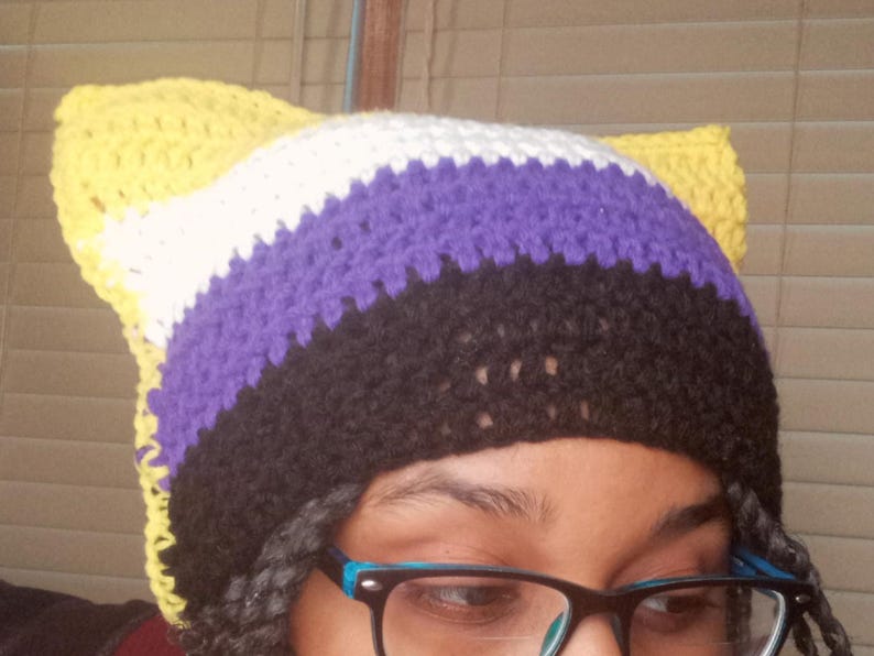 May include: A handmade crochet hat with cat ears, featuring the non-binary pride flag colors: yellow, white, purple, and black. The hat is visible from the forehead up, with the ears standing up. The hat is a unique accessory.