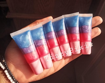 Könnte beinhalten: Sechs Lipgloss-Tuben mit einem Farbverlauf aus Blau-, Lila- und Rosatönen. Die Tuben haben rosa Kappen und werden in einer Hand gehalten. Der Text "TizzyPixie" ist auf die Tuben gedruckt.