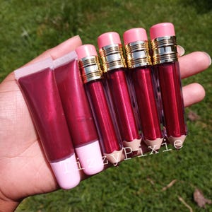May include: A selection of lip gloss products, including tubes and pencil-shaped containers, are held in a hand. The lip gloss is a rich red hue. The pencil-shaped containers have pink lids and gold detailing. The text "Tizzy Pixie" is visible.