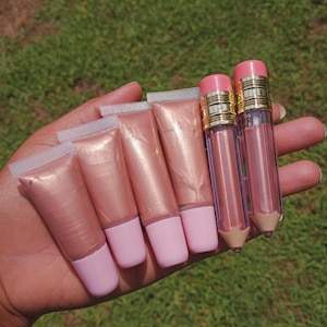 Peut inclure: Une main tenant plusieurs tubes de gloss dans des tons de rose et de pêche. Les tubes de gloss ont des bouchons roses et des tubes transparents. Deux des tubes de gloss ont la forme de crayons avec des accents dorés.