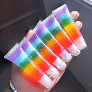 May include: Six clear tubes of rainbow colored lip gloss with a white and purple swirl at the top. The tubes are arranged in a row with the tops of the tubes facing the viewer. The text "Itzy Pixie" is visible on the tubes.
