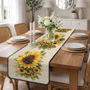 May include: A rectangular beige table runner with a black border, featuring a large sunflower design. The runner is placed on a wooden table set for a meal, with plates, candles, and a vase of flowers.