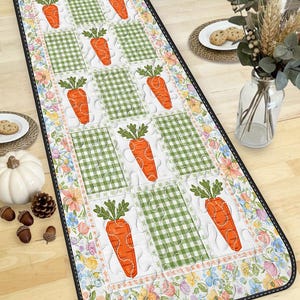 May include: A rectangular table runner featuring a repeating pattern of orange carrots with green tops on white squares, alternating with green gingham squares. The border has a floral design with a black stitched edge. This runner is perfect for Easter or spring decor.