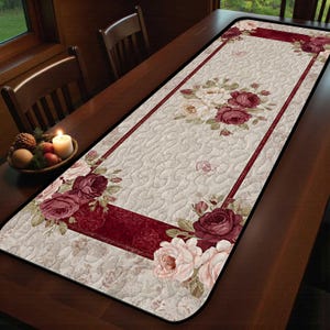 May include: Cream-colored table runner with a floral rose design. The runner features a quilted pattern and a deep red border. Clusters of red and pink roses are arranged along the edges and center, creating a decorative accent for dining tables.
