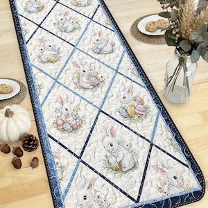 Può includere: Runner da tavolo rettangolare a tema pasquale con un motivo ripetuto di coniglietti bianchi e uova di Pasqua. Il runner ha uno sfondo bianco con un motivo a griglia di diamanti blu e un bordo blu scuro.