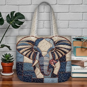May include: A quilted tote bag featuring an elephant design. The elephant is rendered in shades of blue, beige, and brown, with a patchwork effect. The bag has cream-colored handles and a matching background. The elephant is holding a red flower.