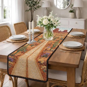 May include: A desert-themed table runner with a detailed design featuring a road, cacti, and desert landscape. The runner is bordered with a black trim and is placed on a wooden table set for a meal. The table is set with white plates, candles, and a floral centerpiece.