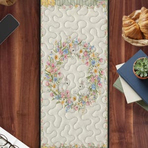 May include: A long, rectangular quilted table runner featuring a central floral wreath with three white bunnies. The runner has a cream background with a wavy pattern, and floral and striped patterned borders in yellow, green, and floral prints.