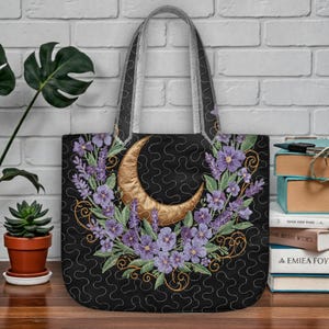 Puede incluir: Bolso tote negro con una luna creciente dorada y una corona de flores moradas. El bolso tiene un diseño acolchado y correas grises. La corona floral incluye lavanda y otras flores moradas con hojas verdes.