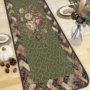 May include: Green floral table runner with a central floral design in pink, cream, and burgundy. The runner has a quilted appearance with a decorative border and a black edge. The runner is approximately 60 inches long.