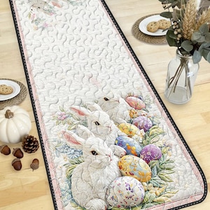 May include: Easter-themed runner featuring white rabbits and colorful Easter eggs. The runner has a quilted design with a pink border and black trim. The design includes floral accents, creating a festive and decorative table runner.