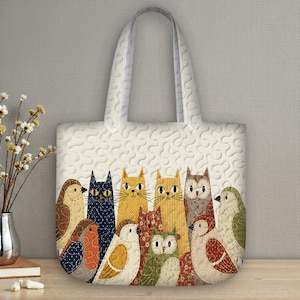 Puede incluir: Bolso tote blanco con un patrón repetitivo y un diseño de patchwork colorido de gatos, pájaros y búhos. El bolso tiene una textura acolchada y asas resistentes, perfecto para llevar comestibles o libros.