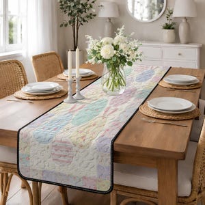 Puede incluir: Camino de mesa pastel de Pascua con un diseño acolchado de huevos y conejos. El camino de mesa está sobre una mesa de comedor de madera con platos blancos, manteles individuales tejidos y un centro de mesa floral. Mide aproximadamente 183 cm de largo.