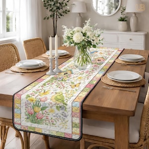 May include: A floral table runner with a bird and butterfly design. The runner features a white background with colorful flowers, birds, and butterflies. The edges are bordered with a black trim and a patchwork design. The runner is placed on a wooden table.