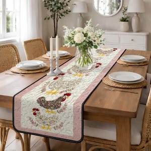 以下が含まれることがあります： クリーム色の背景に、花の鶏の繰り返し模様が施されたキルトテーブルランナー。 ランナーはピンクの縁と黒のトリムがあり、ダイニングテーブルに彩りを添えています。 ランナーは木製のテーブルの上に置かれています。