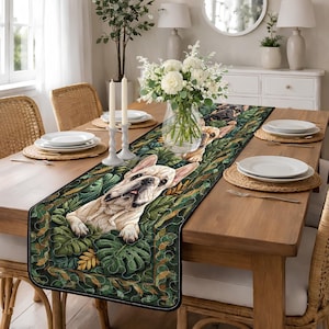 May include: A rectangular table runner featuring a French bulldog design amidst lush green foliage. The runner's border is black with a white stitched edge. The table is set with white plates, woven placemats, and candles, creating a warm, inviting dining scene.