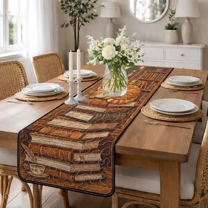 Könnte beinhalten: Ein rechteckiger Tischläufer mit einem Design aus Büchern und einer schlafenden orangefarbenen Katze. Der Läufer ist braun mit einem schwarzen Rand und liegt auf einem Holztisch. Weiße Teller, Kerzen und eine Blumenvase sind ebenfalls vorhanden.