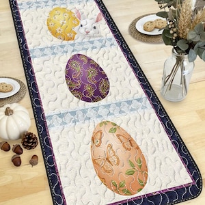 以下が含まれることがあります： 刺繍されたイースターエッグと花柄が特徴の、キルト加工されたイースターテーブルランナー。 ランナーは白地で、青と紫の縁取りがあります。 白いウサギが黄色い卵の後ろから顔をのぞかせています。 長さ約150cm。