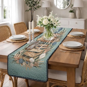 Puede incluir: Un camino de mesa rectangular turquesa con un diseño náutico que presenta conchas marinas e hipocampos. El camino de mesa está colocado sobre una mesa de comedor de madera con platos blancos, manteles individuales tejidos y sillas de mimbre. Un jarrón de flores blancas y velas completan la escena.