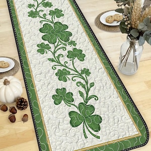 May include: A rectangular table runner with a white background and a green border. The runner features a repeating pattern of green shamrocks and vines. The border is green with a gold trim and black stitching.