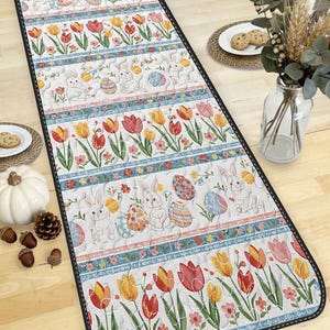 Puede incluir: Camino de mesa con temática de Pascua con un patrón repetido de conejitos blancos, huevos de Pascua decorados y tulipanes coloridos. El camino de mesa tiene un fondo blanco con bordes azules y florales y un borde cosido negro. Mide unos 152 cm de largo.
