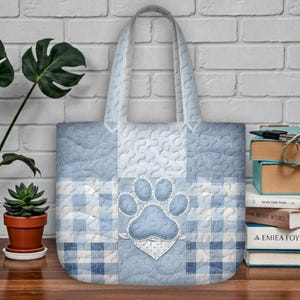 Può includere: Borsa tote trapuntata nei toni del blu e del bianco, con un'applicazione a forma di zampa. La borsa ha un design patchwork con un mix di motivi, tra cui quadretti e texture a vortice. I manici sono azzurri. Tema: animali domestici.