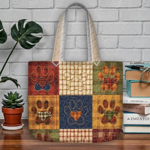 Può includere: Una borsa tote patchwork con impronte di zampe in vari colori e motivi. La borsa ha un manico color crema ed è realizzata in tessuto trapuntato. Il design include quadrati rossi, verdi e gialli con accenti a quadri e tinta unita.