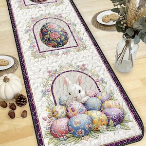 May include: Easter-themed table runner with a white quilted background and a purple border. The runner features floral egg designs and a central image of two white rabbits surrounded by decorated Easter eggs. A decorative piece for the holiday.