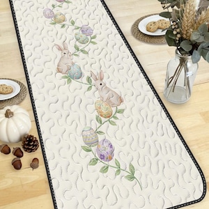 May include: Cream-colored Easter table runner with a black border. The runner features a repeating pattern of bunnies, Easter eggs, and green vines. The eggs are decorated in pastel colors. The runner is placed on a wooden table.