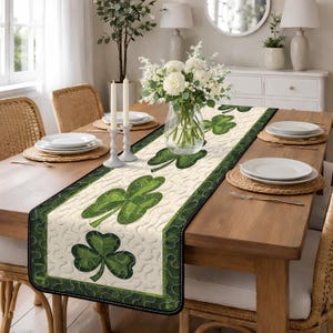Puede incluir: Camino de mesa crema con un diseño de tréboles verdes. El camino de mesa tiene un borde verde oscuro y está colocado sobre una mesa de madera preparada para una comida. También hay platos blancos, velas y un arreglo floral.