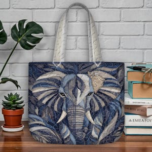 Puede incluir: Un bolso tote azul con un diseño detallado de elefante. El elefante está representado en tonos azules, grises y blancos, con costuras intrincadas. El bolso tiene una correa de color claro y está sobre un fondo de pared de ladrillo.
