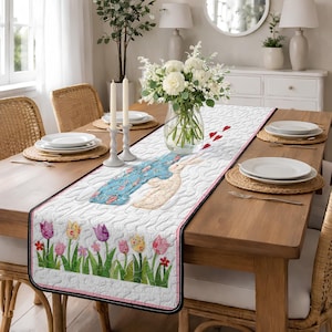 May include: A white quilted table runner featuring a whimsical design of a rabbit with a blue patterned scarf and a tulip border. The runner is placed on a wooden table set for a meal, with plates, candles, and a floral centerpiece.