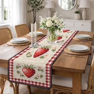 May include: A quilted table runner featuring a strawberry design. The runner has a cream-colored center with large red strawberries and floral accents. A red and white gingham border frames the design. The runner is placed on a wooden table.