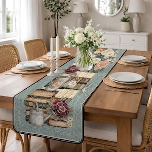 May include: A rectangular table runner with a floral and book design. The runner features a light blue background with a black border. The design includes stacks of books, roses, and a watering can. The table is set with white plates, candles, and a vase of flowers.