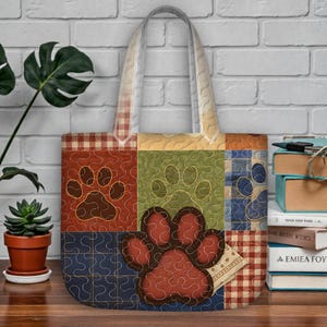 May include: A patchwork tote bag with a large paw print design. The bag features a variety of colours and patterns, including red, green, blue, and tan squares. The paw prints are embroidered with a contrasting thread. The bag has a beige handle.